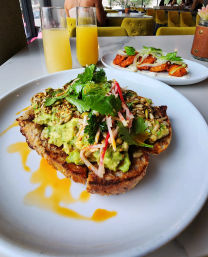 Avocado toast on grilled sourdough topped with smashed avocado, seeds, radish slaw and cilantro, drizzled with sauce, paired with two orange juice flutes and a plate of spicy fried chicken bites on a marble table in a bright café with yellow chairs.