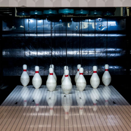 Ten white bowling pins with red neck stripes set on a glossy lane under an automatic pinsetter at an indoor bowling alley, ready for a strike