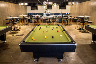 Modern indoor entertainment lounge with a green pool table in the foreground, bowling lanes and high-top seating with gold stools, pendant lights and wood-paneled walls.