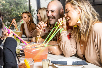 Group of friends laughing at a restaurant bar, sharing a large pink tropical punch bowl with bright neon curly straws and cocktails on the table