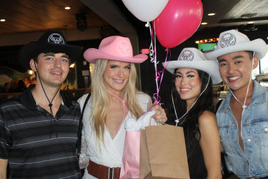 Four smiling friends in cowboy hats at a bar celebration holding pink and white balloons and gift bags