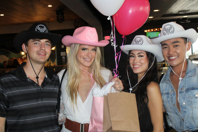 Four smiling friends in cowboy hats at a bar celebration holding pink and white balloons and gift bags