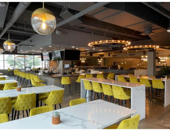 Cheerful urban restaurant interior with marble-topped communal tables, tufted yellow-green barstools and chairs, circular Edison-style pendant chandeliers, central bar and large windows.