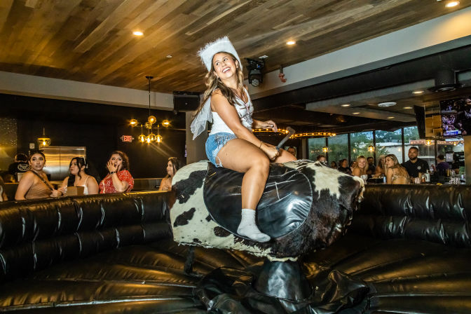 Smiling woman in a white cowboy hat and denim shorts rides a cow-pattern mechanical bull indoors while a crowd of onlookers watches in a lively bar setting
