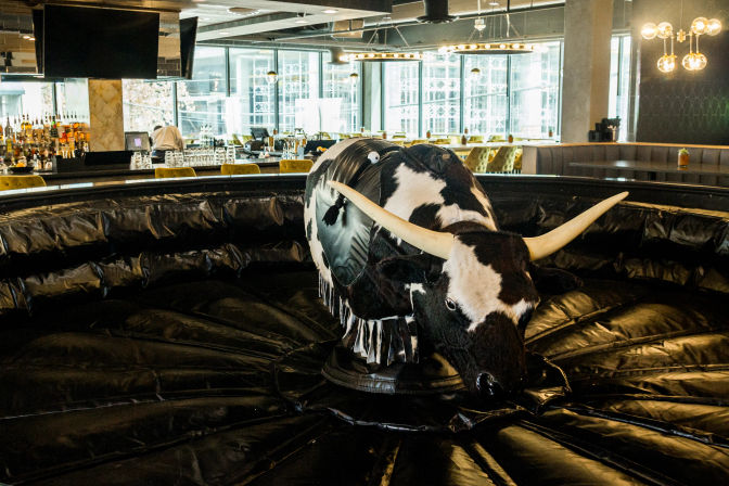 Mechanical bull with black-and-white cow pattern and long horns on a circular inflatable mat inside a modern indoor bar and lounge with yellow barstools and large windows