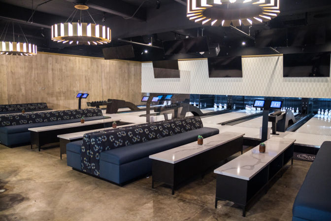 Modern indoor bowling alley interior with polished lanes and pins, digital scoring screens, patterned blue banquette seating, marble-top tables and large circular pendant lights