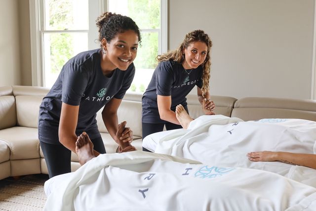 Two smiling therapists giving in-home foot massages to clients covered in white sheets in a bright living room — mobile spa couples massage and relaxation service