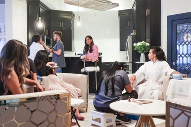 Group of women enjoying a home spa day in a modern open-plan kitchen and living room — a woman in a white robe gets a manicure while others chat, sip drinks, and relax near black cabinets, pendant lights, and fresh flowers.