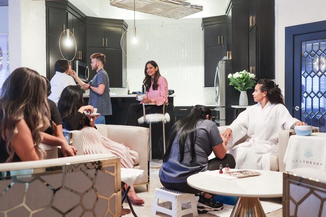 Group of women enjoying a home spa day in a modern open-plan kitchen and living room — a woman in a white robe gets a manicure while others chat, sip drinks, and relax near black cabinets, pendant lights, and fresh flowers.