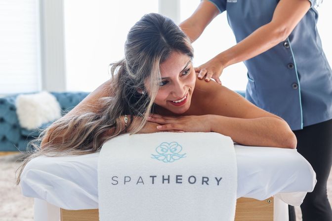 Smiling woman lying on a massage table with a white towel, receiving a relaxing back massage from a therapist in a bright, airy spa room.