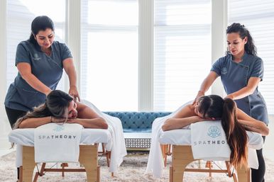 Bright spa treatment room with two clients receiving side-by-side back massages from therapists on white linen-covered massage tables, soft rug and tufted blue sofa in the sunlit background.