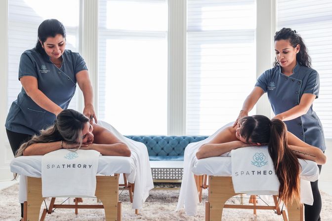 Bright spa treatment room with two clients receiving side-by-side back massages from therapists on white linen-covered massage tables, soft rug and tufted blue sofa in the sunlit background.