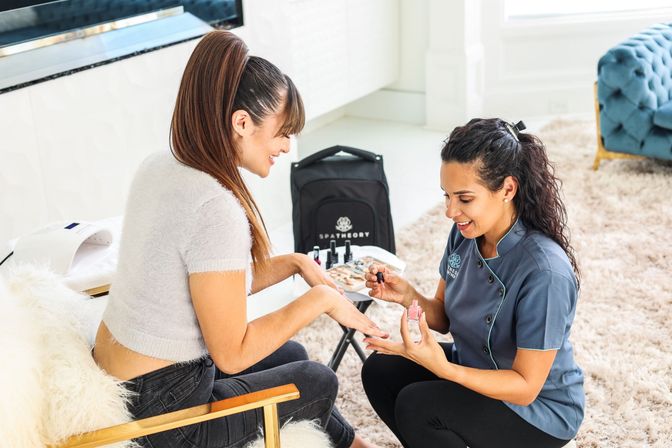 Smiling nail technician giving an at-home manicure, painting a client's nails with pink polish in a bright modern living room with a plush rug and blue tufted sofa