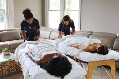 Two therapists giving foot massages to two clients on portable massage tables in a bright living-room in-home spa scene