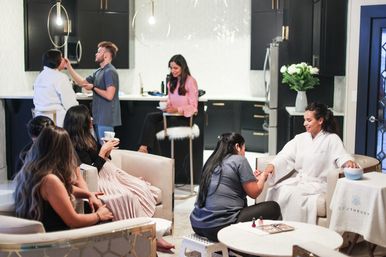 Women enjoying a pampering spa day in a modern lounge — a guest in a white robe gets a manicure while another has makeup applied and others sip tea and relax in a bright, stylish space.