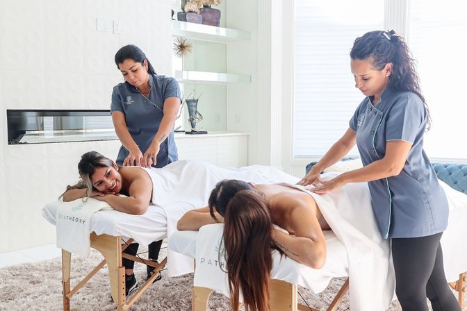 Two women enjoying side-by-side back massages from therapists in matching gray uniforms in a bright, modern spa room with white linens and a plush rug.