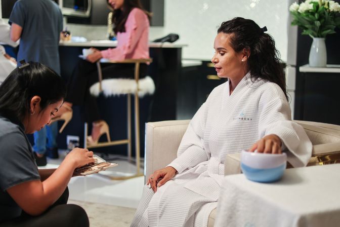 Client in a white spa robe relaxing in a modern day-spa lounge with her hand in a bowl while a nail technician prepares tools for a manicure.