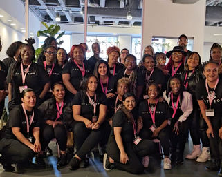 Large, smiling group of women and a few men wearing matching black shirts and pink lanyards posing for a team photo inside a bright, modern coworking/event space.