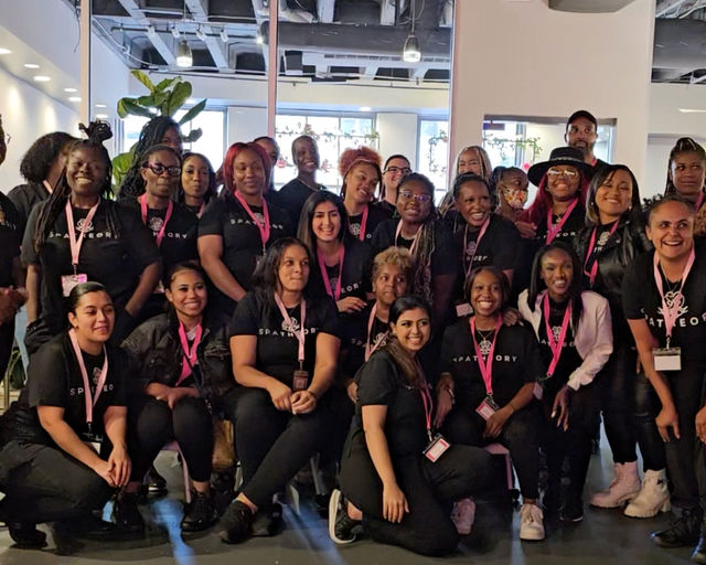 Large, smiling group of women and a few men wearing matching black shirts and pink lanyards posing for a team photo inside a bright, modern coworking/event space.