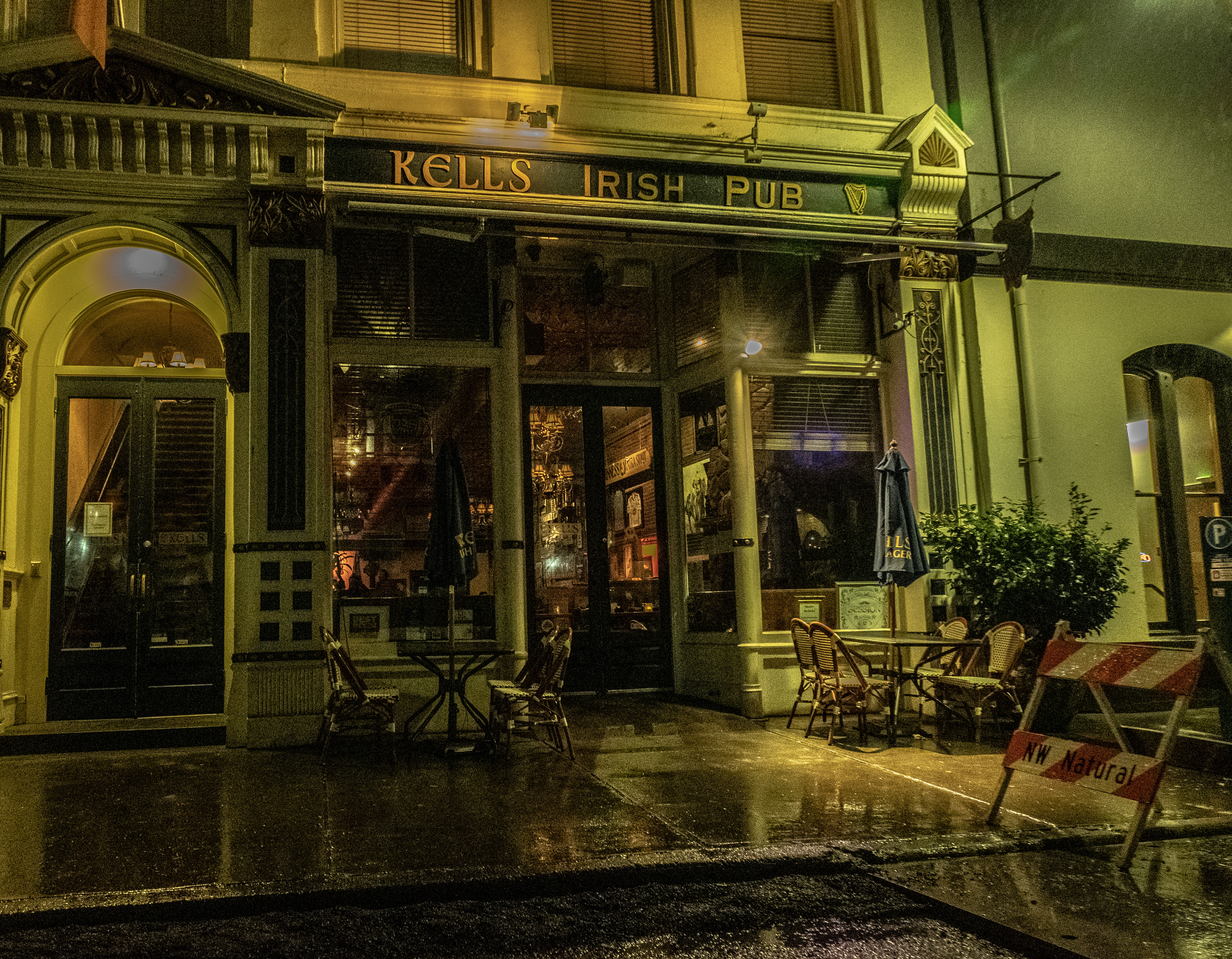 Rainy night street view of an Irish pub storefront with warm interior lights, empty outdoor tables and wicker chairs under closed umbrellas, ornate historic facade, glossy wet pavement reflecting light, and a sidewalk construction barricade.