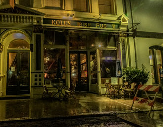 Rainy night street view of an Irish pub storefront with warm interior lights, empty outdoor tables and wicker chairs under closed umbrellas, ornate historic facade, glossy wet pavement reflecting light, and a sidewalk construction barricade.
