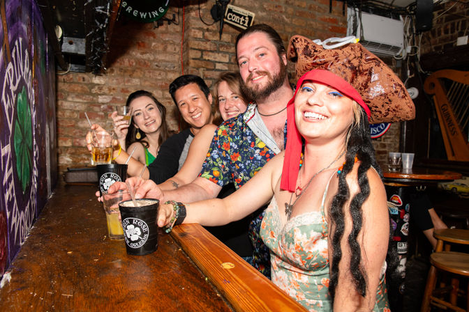 Five friends smiling at a brick‑walled New Orleans bar counter, woman in front wearing a playful pirate hat and floral dress raising a drink, others holding cocktails for a festive night out
