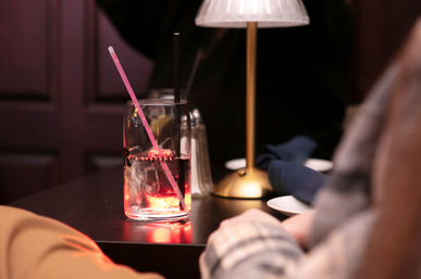 Close-up of a glass with ice and a pink straw glowing on a dim restaurant table under a warm lamp, with napkin and plates nearby