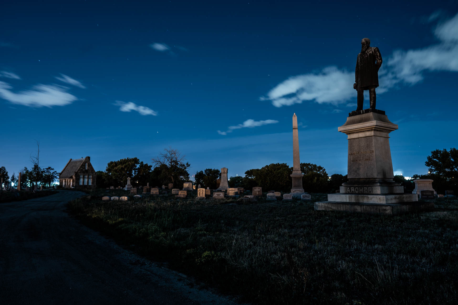 Moonlit historic cemetery at night with a tall statue on a pedestal, a slender obelisk, rows of stone grave markers and a small stone chapel under a deep blue starry sky.