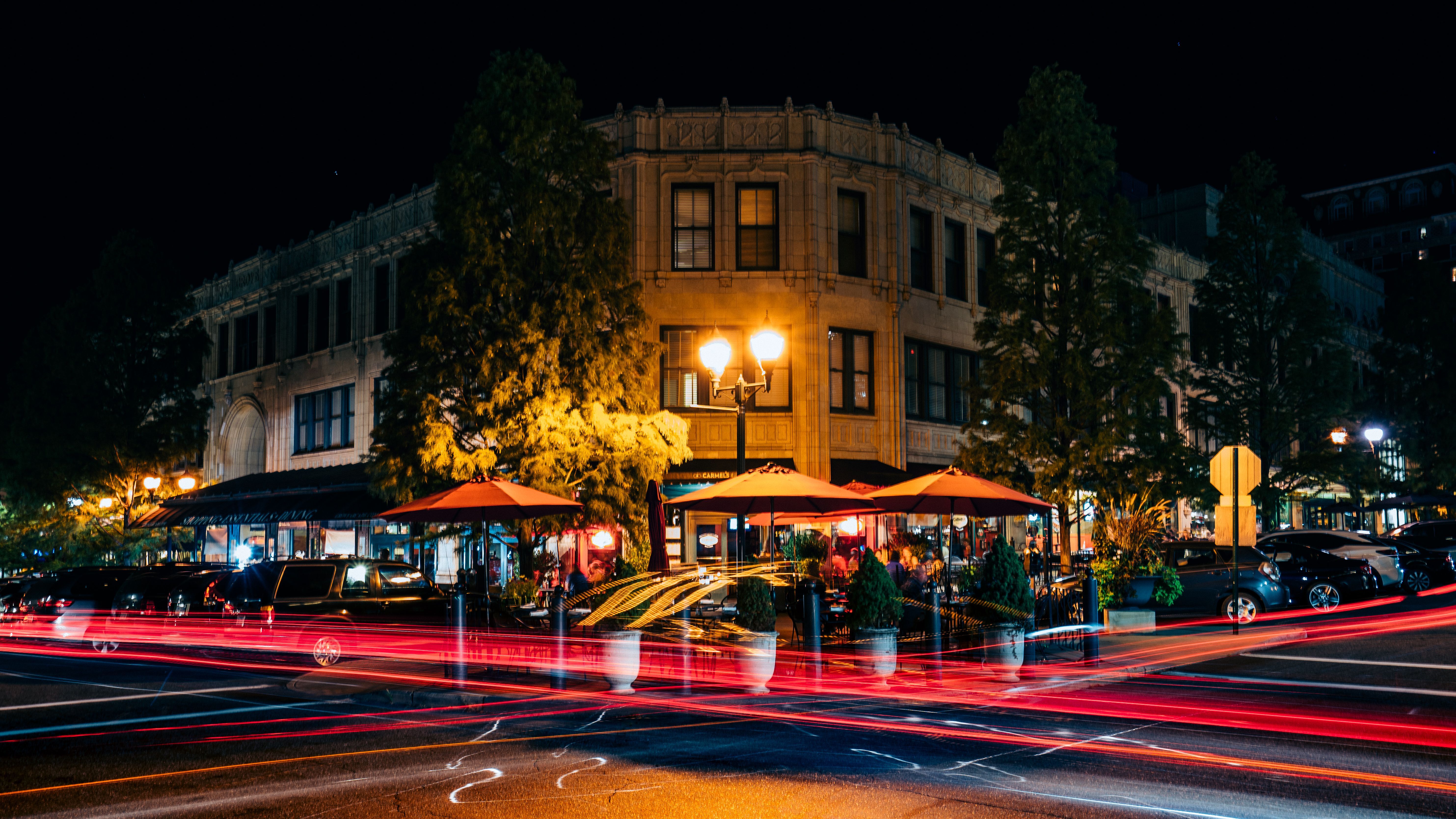 Vibrant downtown night scene: historic corner building with lit streetlamps and outdoor restaurant patios under orange umbrellas, framed by trees and streaking red car light trails.