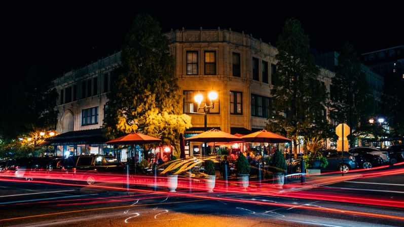 Vibrant downtown night scene: historic corner building with lit streetlamps and outdoor restaurant patios under orange umbrellas, framed by trees and streaking red car light trails.