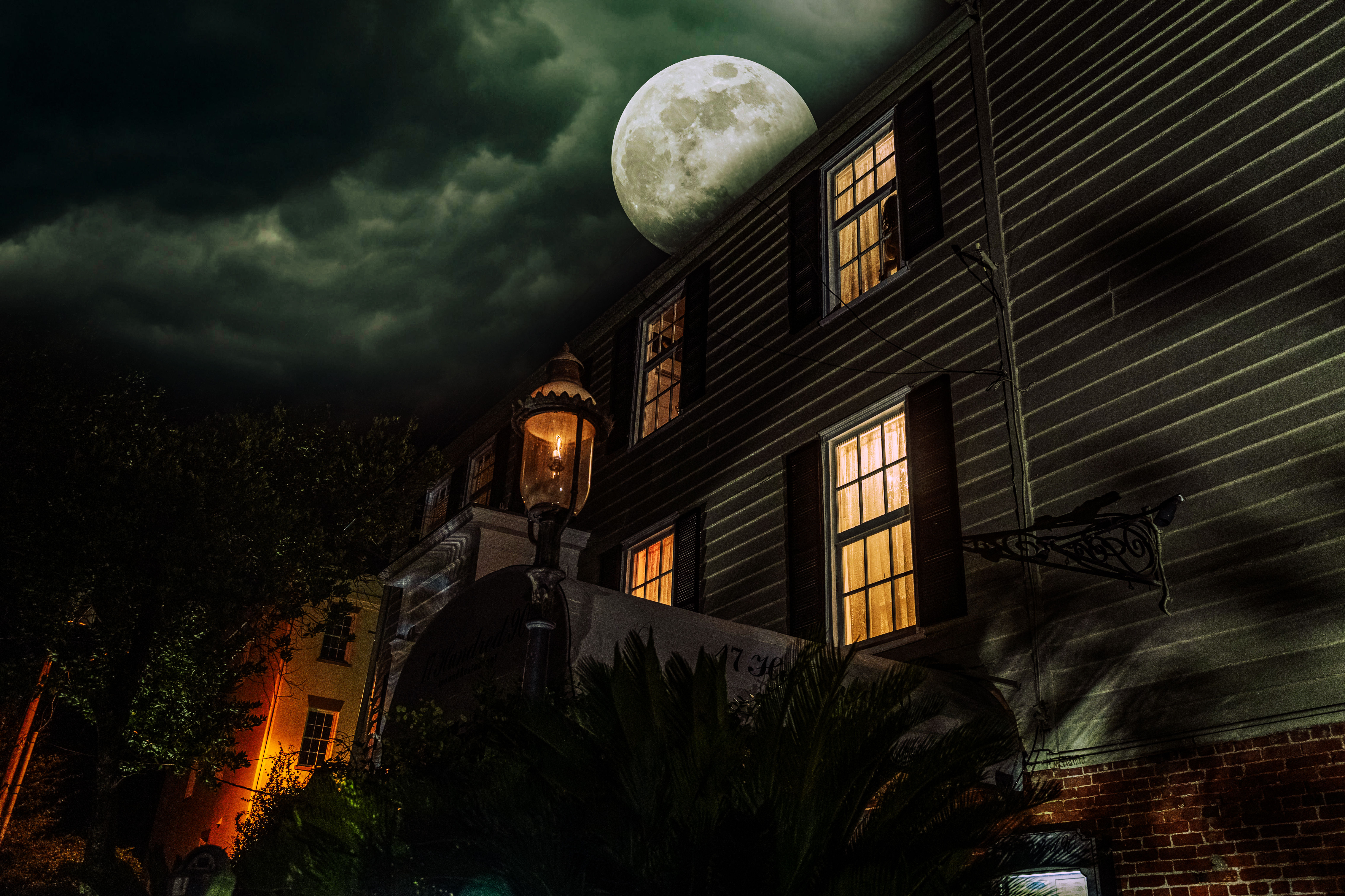 Moonlit historic house exterior at night with warm glowing windows, a vintage streetlamp, dramatic cloudy sky and a looming full moon
