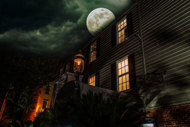 Moonlit historic house exterior at night with warm glowing windows, a vintage streetlamp, dramatic cloudy sky and a looming full moon
