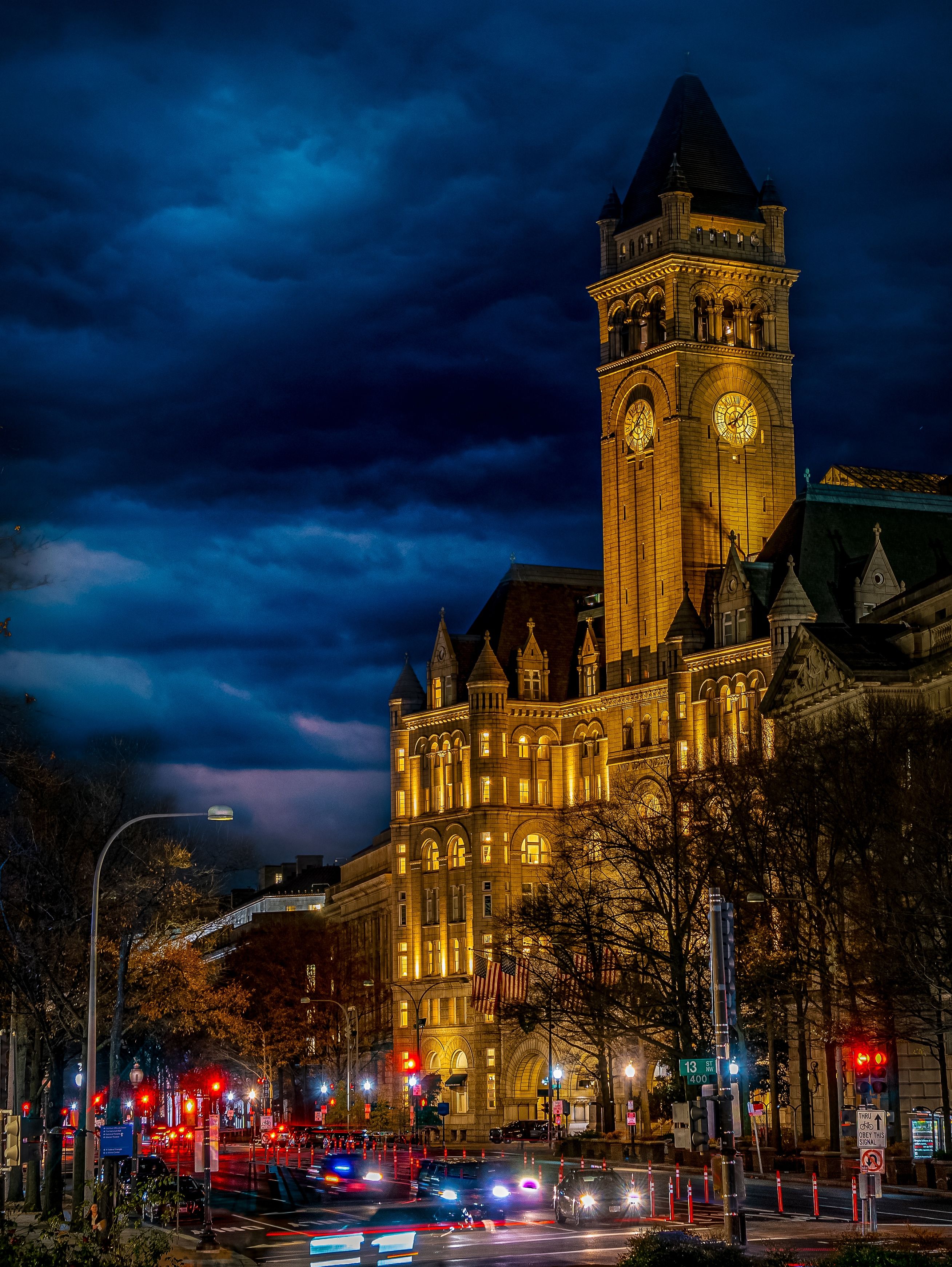 Illuminated historic clock tower and sandstone municipal building at night under a stormy deep-blue sky, downtown street with traffic light trails and glowing streetlights.