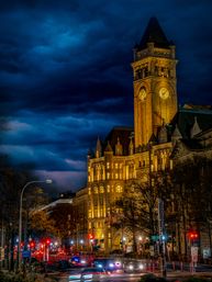 Illuminated historic clock tower and sandstone municipal building at night under a stormy deep-blue sky, downtown street with traffic light trails and glowing streetlights.