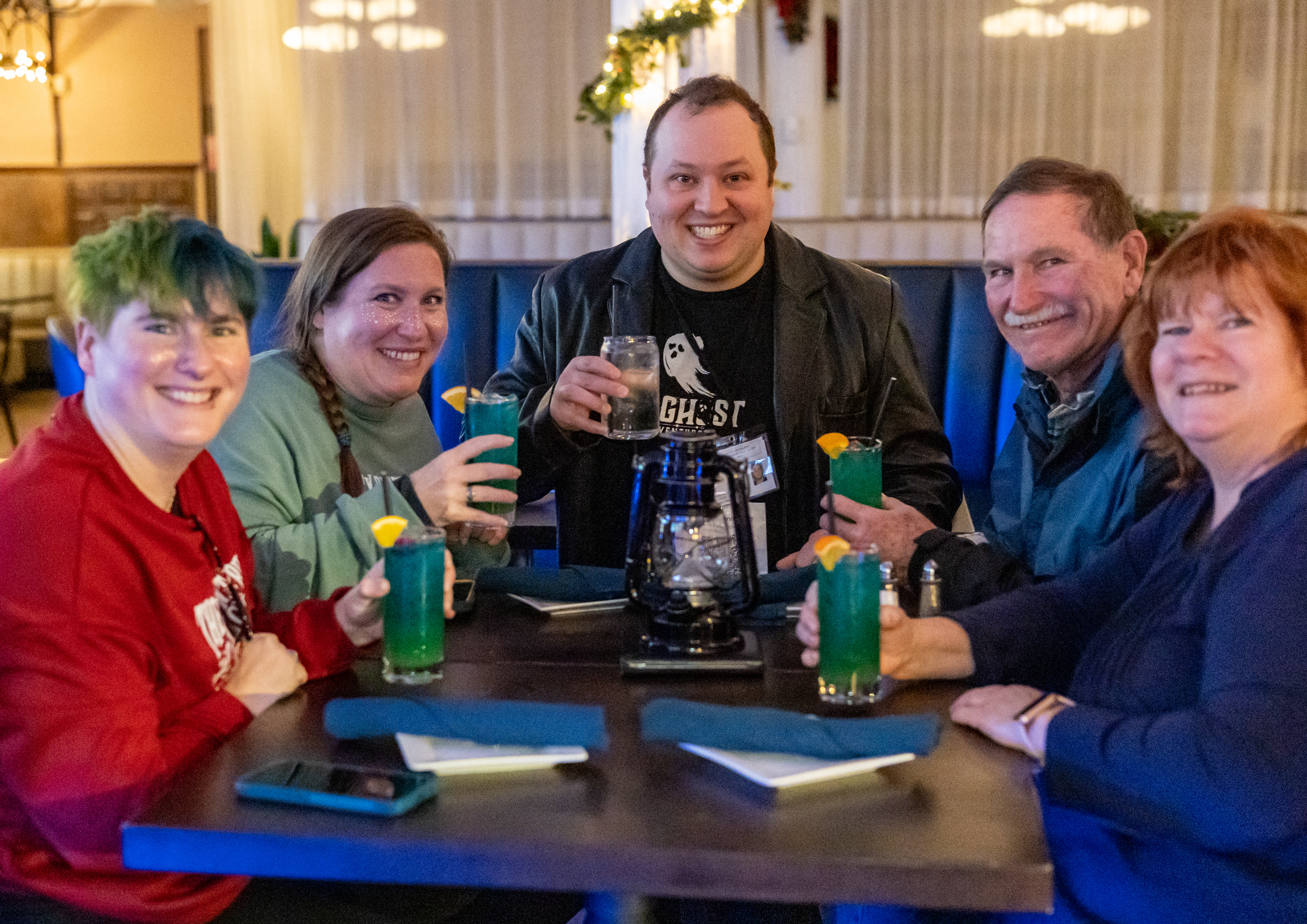 Five smiling adults at a cozy restaurant booth toasting with turquoise cocktails garnished with orange slices and a clear drink, lantern centerpiece and festive garland in warm-lit interior.