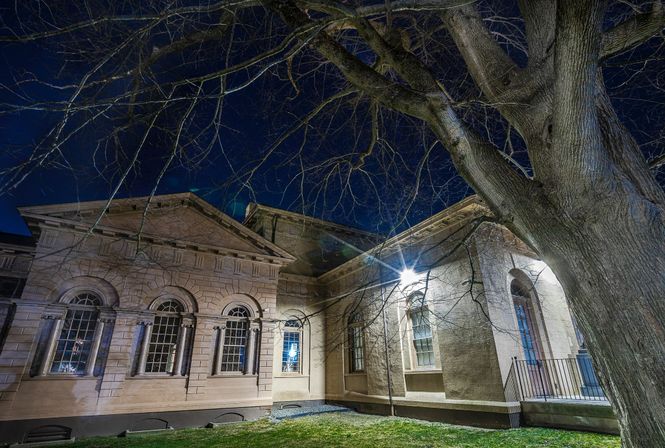 Historic stone civic building at night with arched windows and a bright exterior light, bare tree branches in the foreground under a deep blue sky