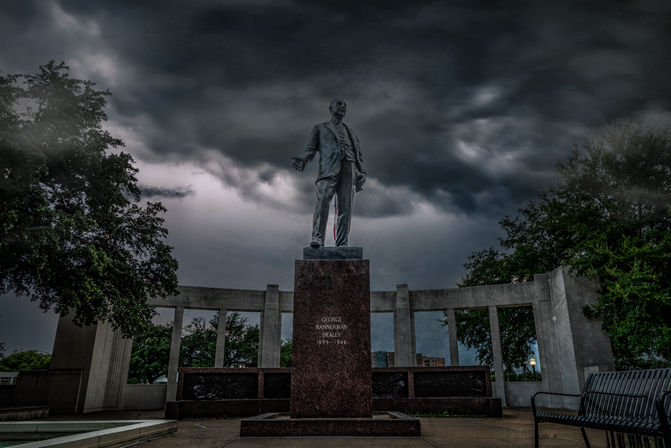 Bronze statue on a granite pedestal in an urban memorial plaza with a curved colonnade, empty bench, trees and dramatic storm clouds overhead