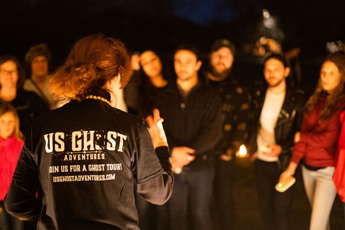 Person leading an outdoor nighttime ghost tour, back to camera in a dark branded jacket, speaking to a small group holding candles and flashlights under warm golden light.