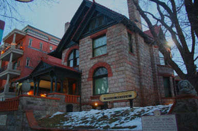 Stone Victorian house museum with arched windows, covered porch and lion statue, snow-dusted lawn and a 'Votes for Women' plaque at dusk