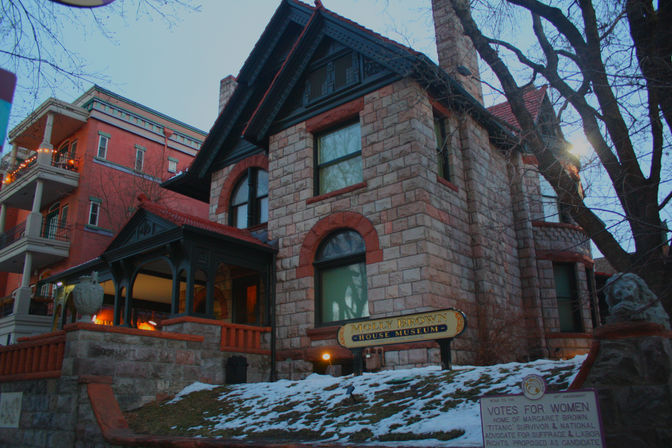 Stone Victorian house museum with arched windows, covered porch and lion statue, snow-dusted lawn and a 'Votes for Women' plaque at dusk