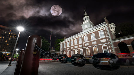Low-angle night view of a historic brick clock-tower building in a downtown plaza, heavy chain and bollards in the foreground, bright streetlamp, red light trails and a dramatic moonlit cloudy sky.