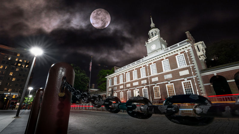 Low-angle night view of a historic brick clock-tower building in a downtown plaza, heavy chain and bollards in the foreground, bright streetlamp, red light trails and a dramatic moonlit cloudy sky.
