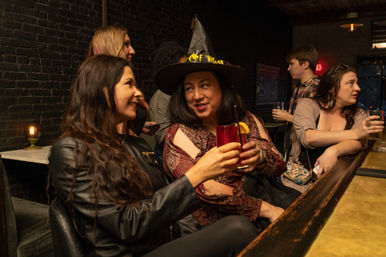 Group of friends toasting at a cozy brick-walled bar — a smiling woman in a witch hat clinks a red cocktail with a friend as others chat at the bar.