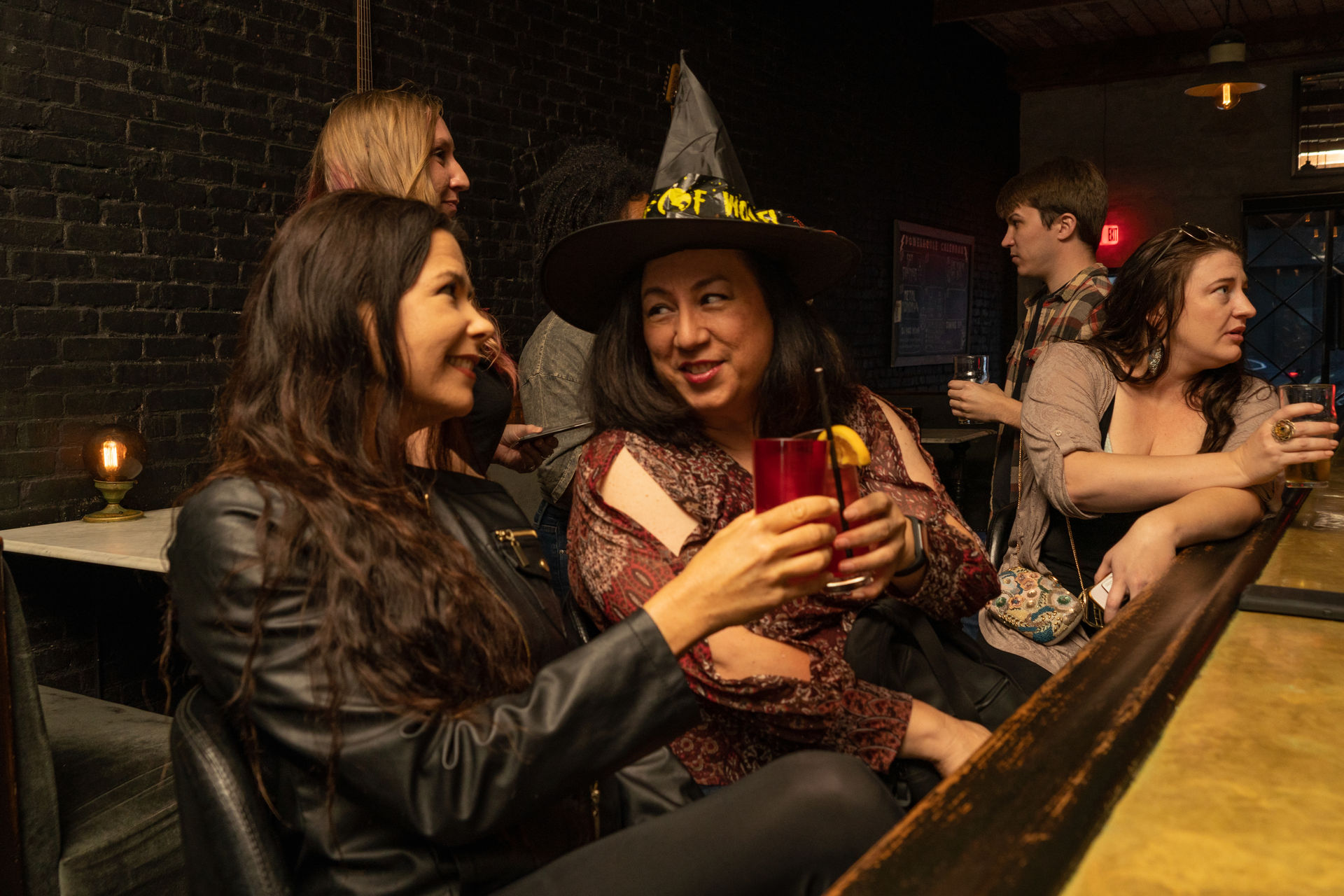 Group of friends toasting at a cozy brick-walled bar — a smiling woman in a witch hat clinks a red cocktail with a friend as others chat at the bar.