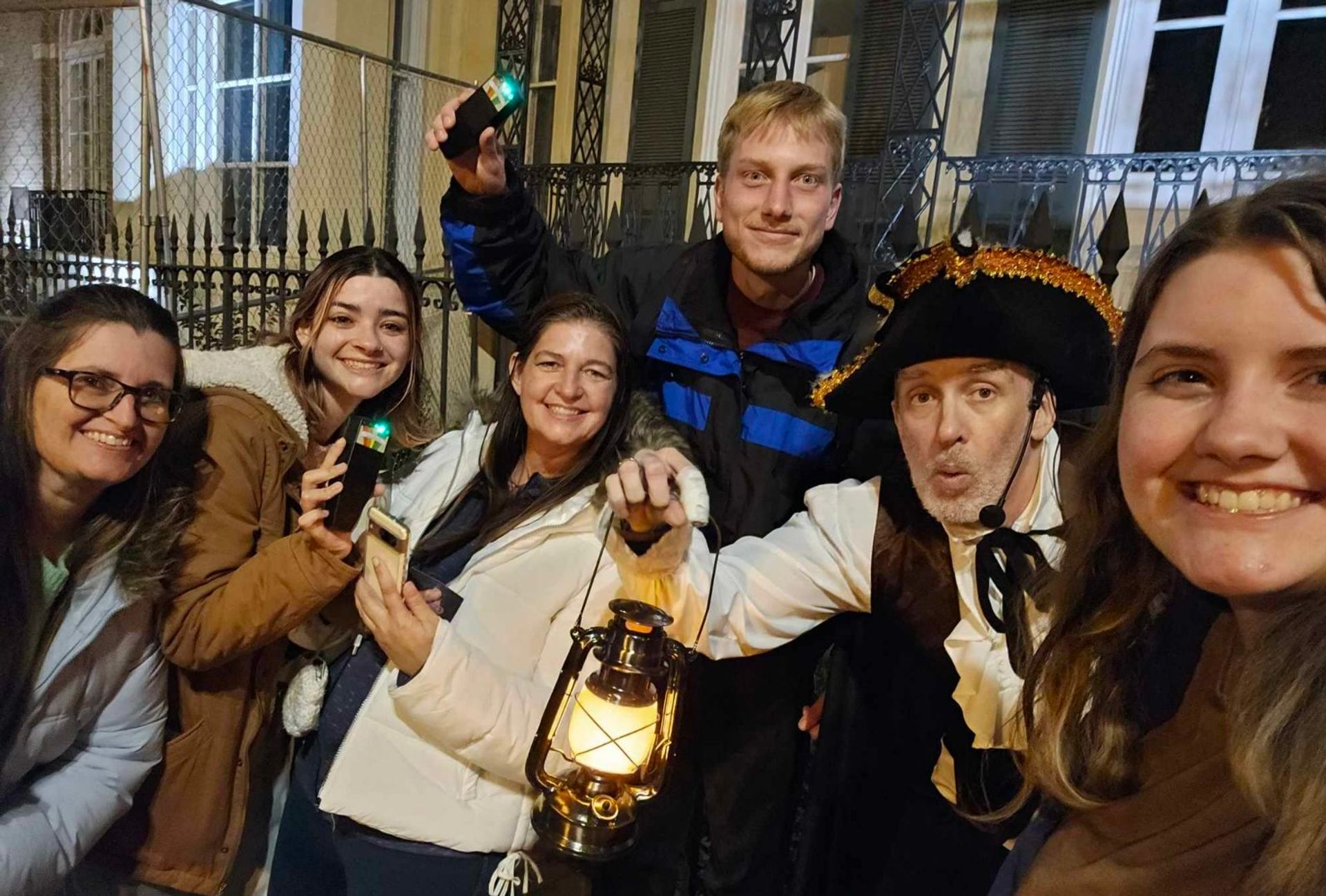 Smiling group selfie on a nighttime historic walking tour in a city street, with a costumed guide wearing a tricorn hat holding a lit lantern and participants holding handheld audio devices with green indicator lights.