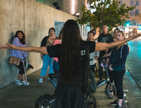 Enthusiastic guide with arms outstretched addressing a small group on a nighttime urban sidewalk near electric scooters, concrete wall, tree, and city streetlights.