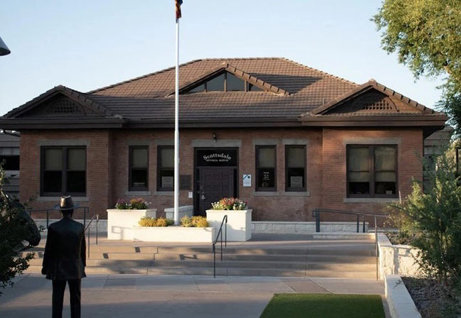 Cozy red-brick Southwestern civic building with a tiled roof, central flagpole, planter steps and a bronze statue facing the entrance on a sunny day.