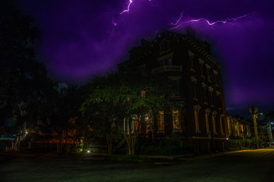 Historic brick corner building on a tree-lined city street at night, warm windows glowing beneath a dramatic purple sky streaked with lightning.