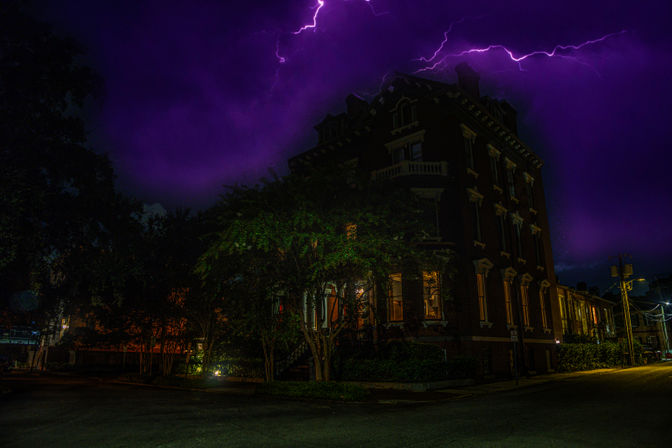 Historic brick corner building on a tree-lined city street at night, warm windows glowing beneath a dramatic purple sky streaked with lightning.