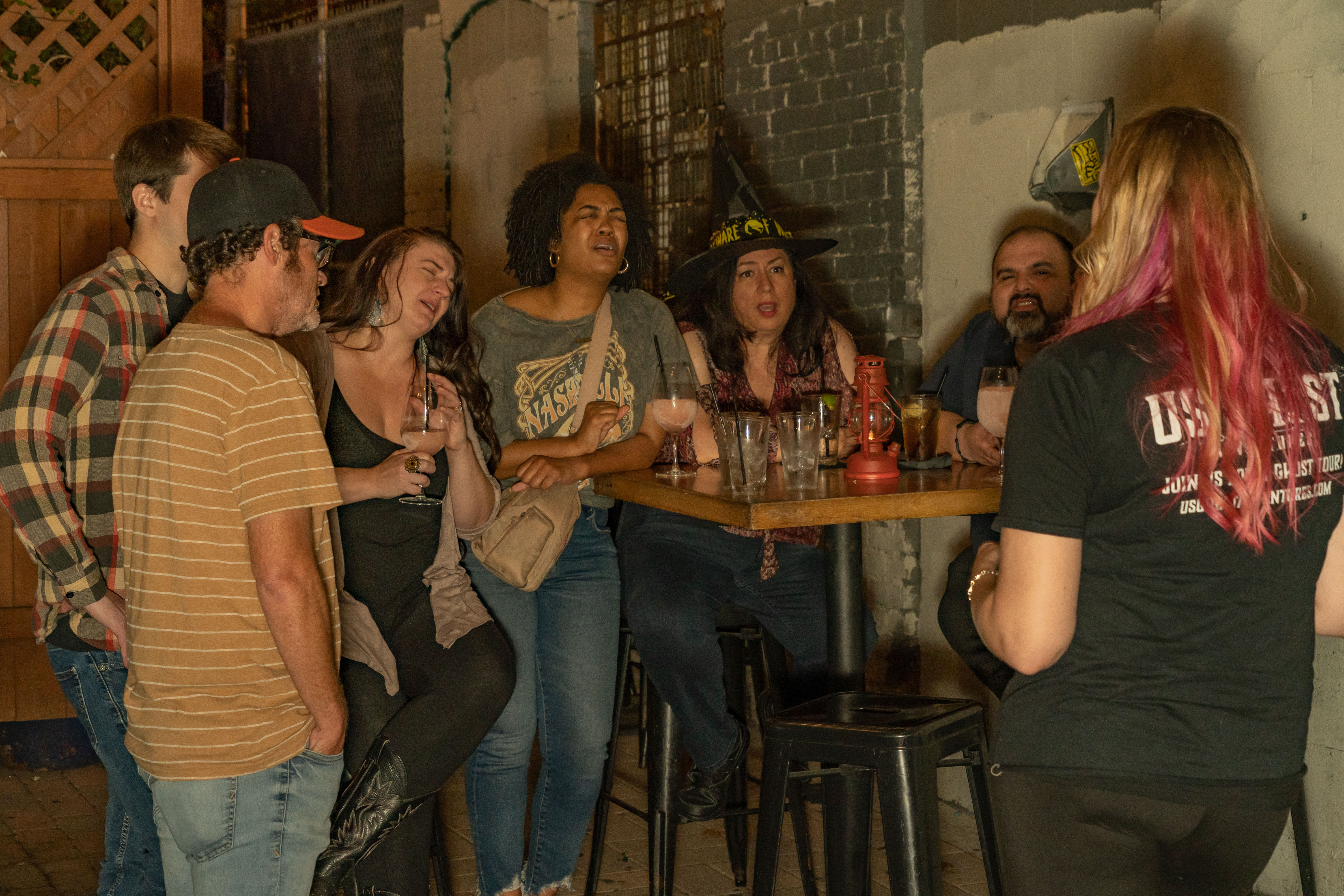 Adults enjoying nightlife at an urban bar patio: a group clustered around a high table with cocktails, one person in a witch hat and a pink‑haired guide addressing them.